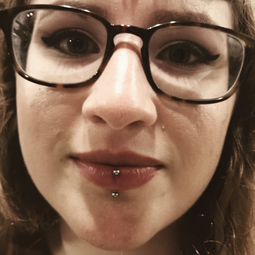 Black and white photo of a person with glasses and text 'Vertical Labret Piercing by Jinx'.