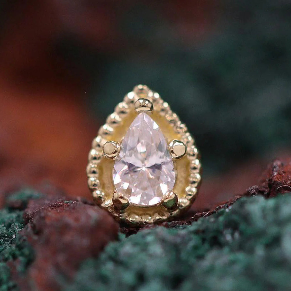 Gold teardrop-shaped earring with a pink gemstone on a textured surface.