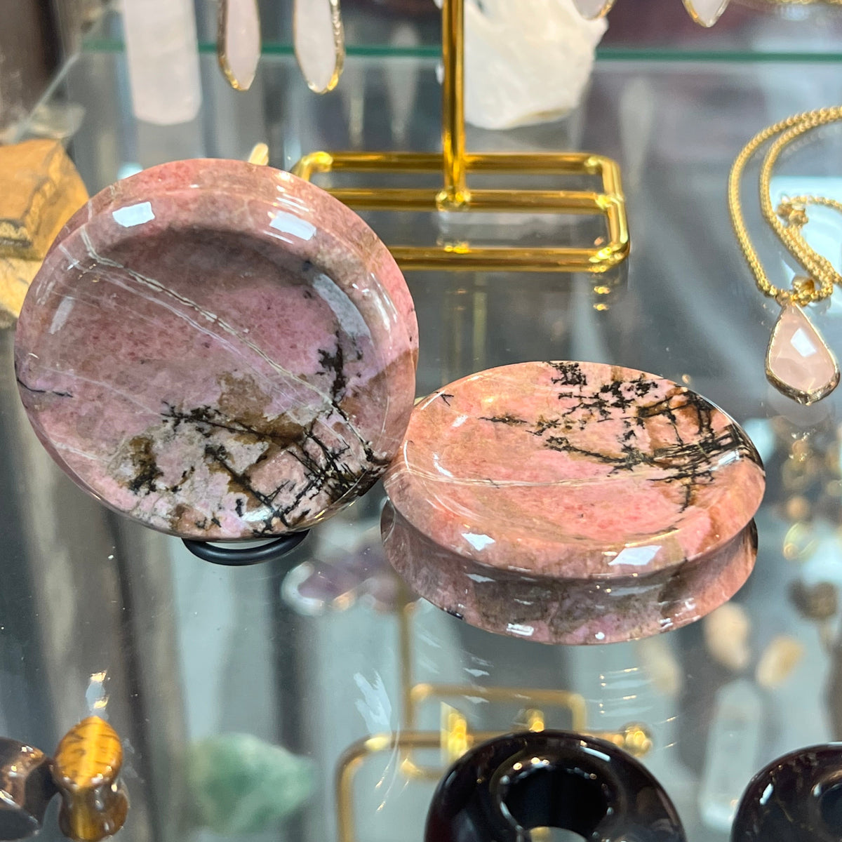 Two round polished pink stone discs with black veining displayed on a glass jewelry stand, with other crystals and jewelry visible in the background.