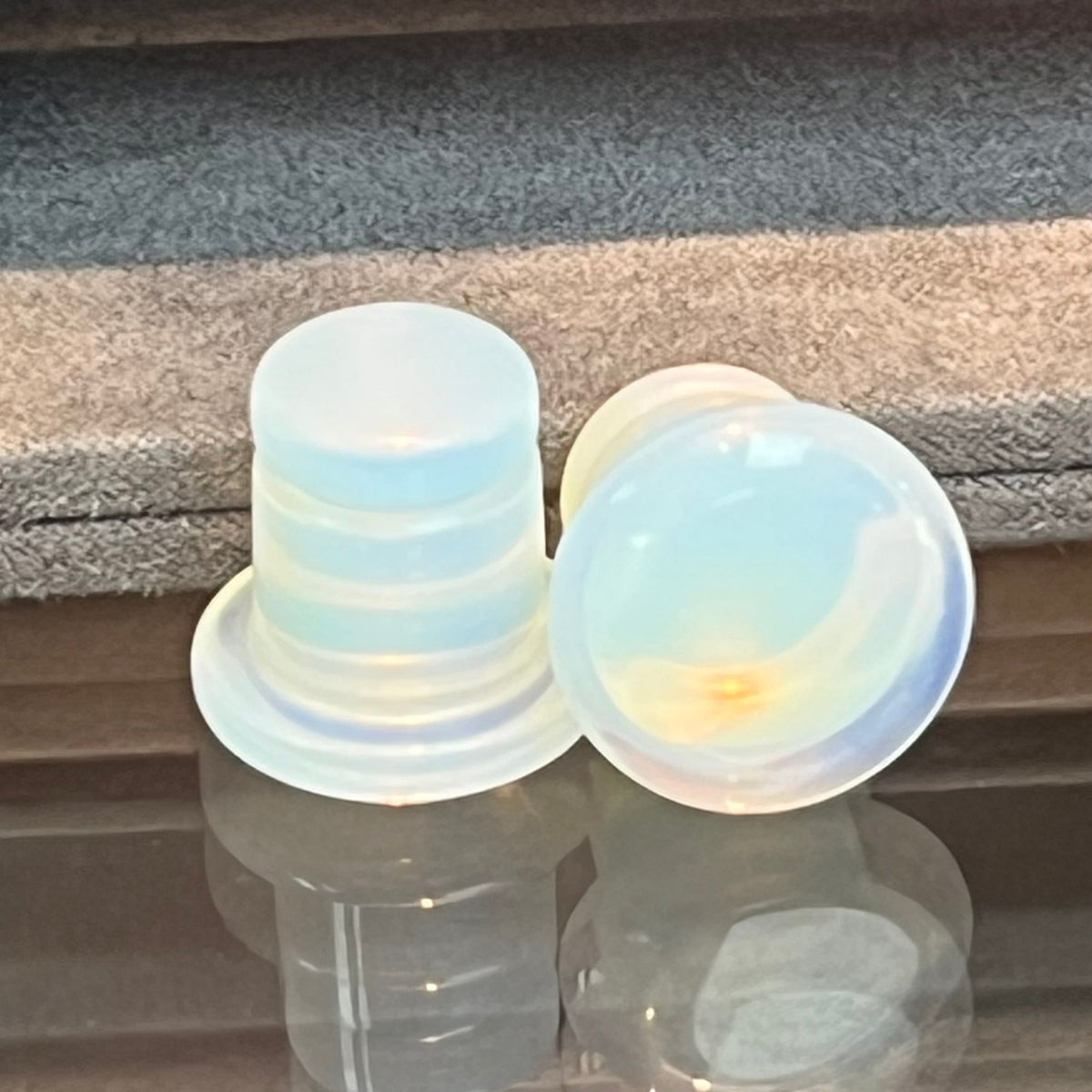A pair of translucent iridescent silicone earplugs resting on a reflective glass surface, with a gray velvet display cushion in the background.