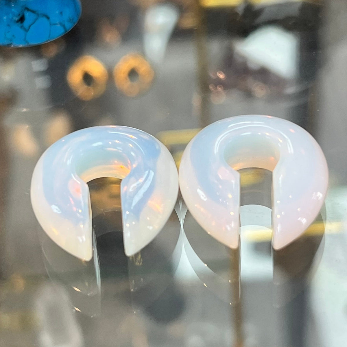 A pair of white crescent-shaped plug earrings displayed on a stand inside a glass jewelry case, with other jewelry pieces and stones visible in the background.