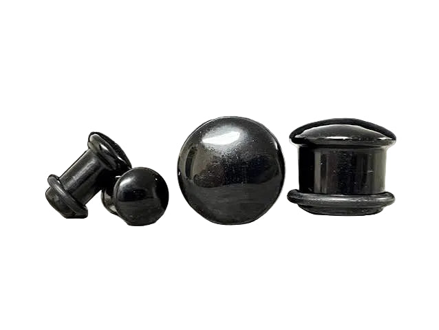 Four black body jewelry pieces, including two flatback plugs and two ball plugs, displayed on a textured gray surface with a blurred gray background.